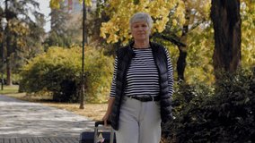 Elderly woman with short grey hair walking with her suitcase along a path in a sunny autumn park - Powered by Shutterstock - Get 15% off with code: PIKWIZARD15