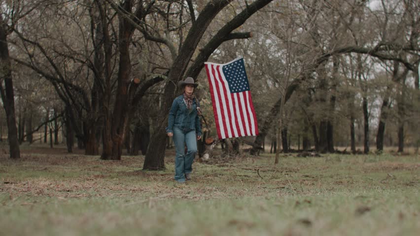 Cowgirl Woman With Braid and Scarf Wearing Cowboy Hat and Denim Jacket Holding Shotgun Looking Ahead With American Flag and Tree in Field With Brown Horses Walking in the Background
