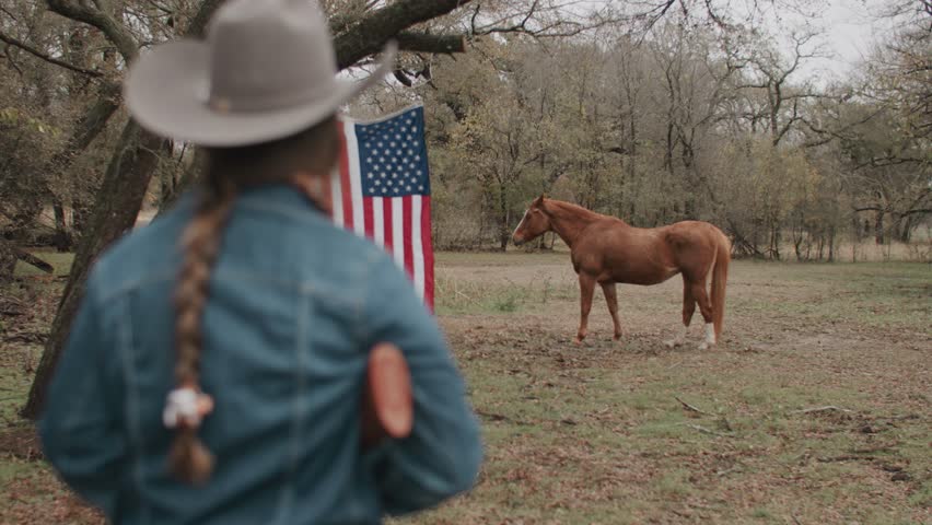 Cowgirl Woman With Braid and Scarf Wearing Cowboy Hat and Denim Jacket Holding Shotgun Looking Ahead With American Flag and Tree in Field With Brown Horses Walking in the Background