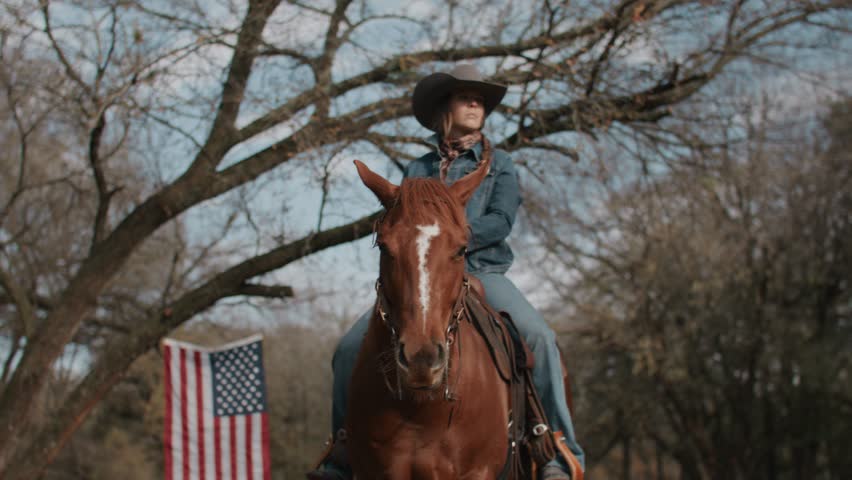 Cowgirl Woman With Braid and Scarf Wearing Cowboy Hat and Denim Jacket Sitting On Brown Horse With American Flag and Tree in Field in the Background