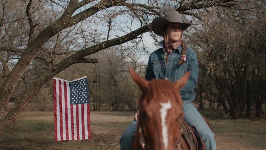 Cowgirl Woman With Braid and Scarf Wearing Cowboy Hat and Denim Jacket Sitting On Brown Horse With American Flag and Tree in Field in the Background