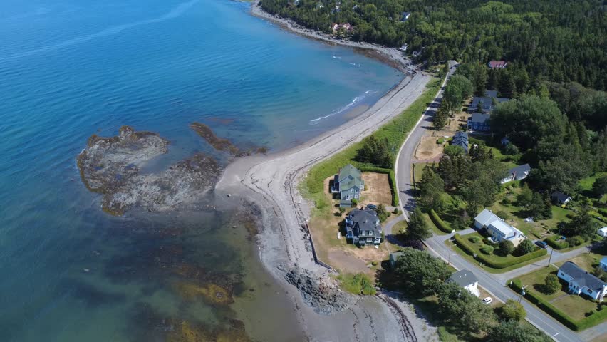 Aerial view of a coastal village bordered by fine sand, rocky reefs with a winding road, green trees and houses along the shore of the St. Lawrence River, Quebec, Canada, 2025.