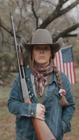 Cowgirl Woman With Braid and Scarf Wearing Cowboy Hat and Denim Jacket Holding Shotgun Looking Off In the Distance With American Flag and Tree in Field in the Background