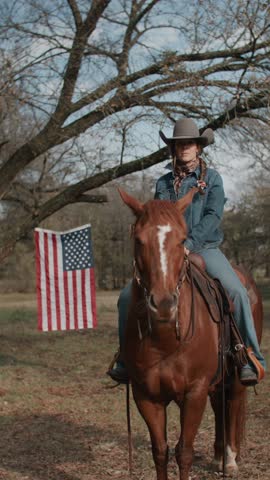 Cowgirl Woman With Braid and Scarf Wearing Cowboy Hat and Denim Jacket Sitting On Brown Horse With American Flag and Tree in Field in the Background