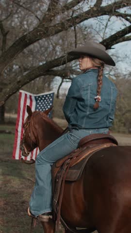 Cowgirl Woman With Braid and Scarf Wearing Cowboy Hat and Denim Jacket Sitting On Brown Horse With American Flag and Tree in Field in the Background