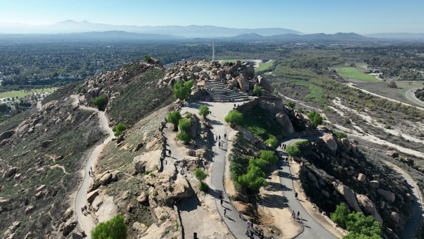 Aerial drone footage of Mount Rubidoux in Riverside, California, showcasing the historic summit cross, memorial stone tower, hiking trails, visitors taking photographs, and panoramic views of the Inland Empire on a sunny day.