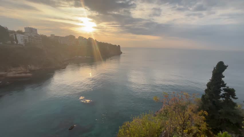 A peaceful panoramic view of the Mediterranean coastline in Antalya, Turkey, showcasing turquoise water, rocky cliffs, and hillside homes overlooking the sea. Captured in warm daylight.