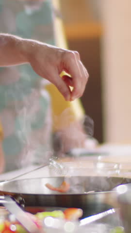 Vertical close-up shot of hand of unrecognizable male chef sprinkling salt to pan during cooking master class