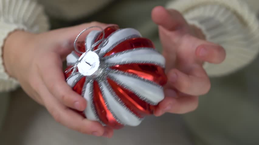 Close-up view of child hands gently holding red white Christmas ornament, warm festive lights creating cozy seasonal atmosphere. Boy swings Christmas red and white ball.