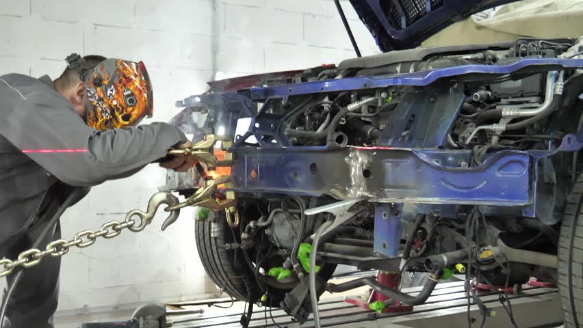 A welder wearing a protective mask welds the front part of a car frame on a repair stand in a workshop
