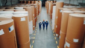 Employees walk past huge paper rolls inspecting, Personnel in helmets assess rolls of paper in aisle, Supervisors and engineers conduct organized equipment checks in industrial warehouse setting - Powered by Shutterstock - Get 15% off with code: PIKWIZARD15