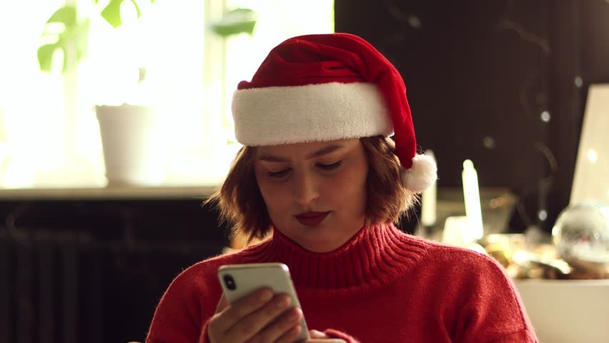 Portrait of happy young woman wearing winter red sweater typing online message using mobile phone on Christmas background with at cozy light living room with festive interior.
