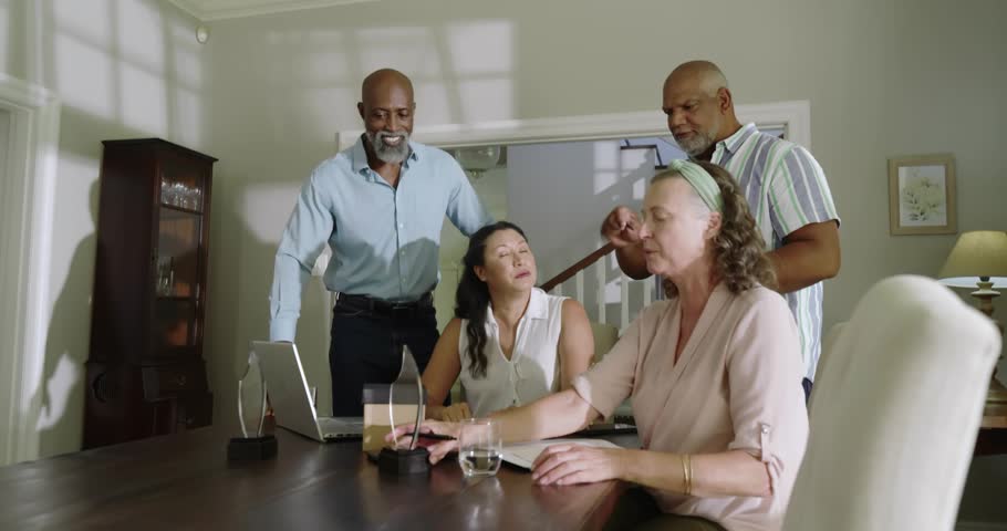 Four adults gathering, man reaching award and causing business globe on laptop checking papers. Dining, home, daylight, lifestyle, collaboration, discussion, teamwork