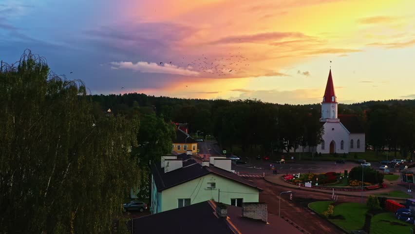 Latvian town Smiltene with church tower under purple-orange sky at sunset with bird flock