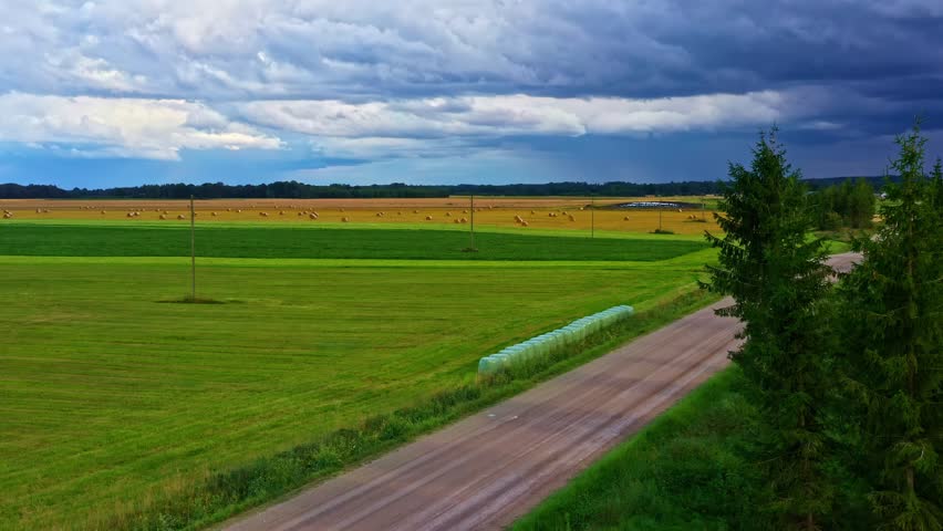 Country road with hay bales and darkening storm clouds over flat farmland in aerial pedestal up
