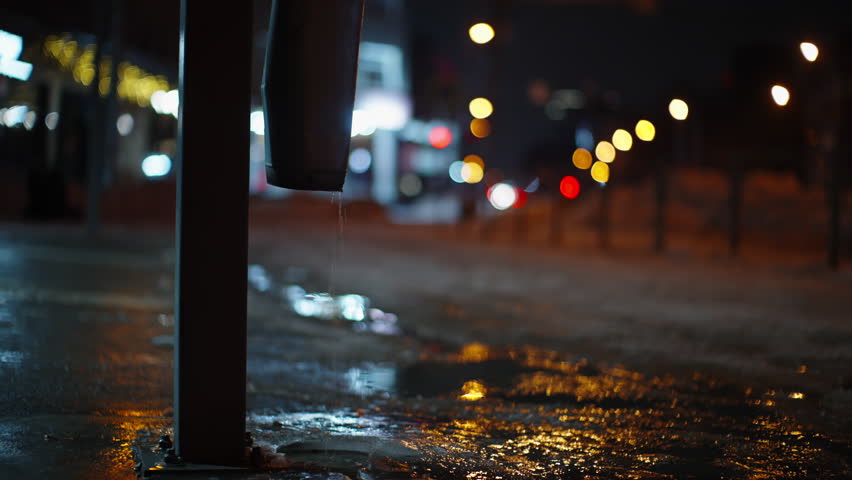 Dripping drainpipe on an icy urban sidewalk at night, forming shimmering puddles that reflect colorful bokeh streetlights, capturing a cold winter city scene with moody atmosphere.
