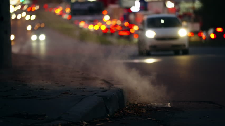 Urban evening traffic with blurred headlights and taillights glowing in the background as steam rises from a city curb vent, capturing the moody atmosphere of a roadway at dusk.