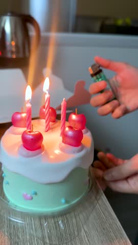Hand lights pink striped candles on pastel cake in kitchen, warm indoor lighting, close-up view