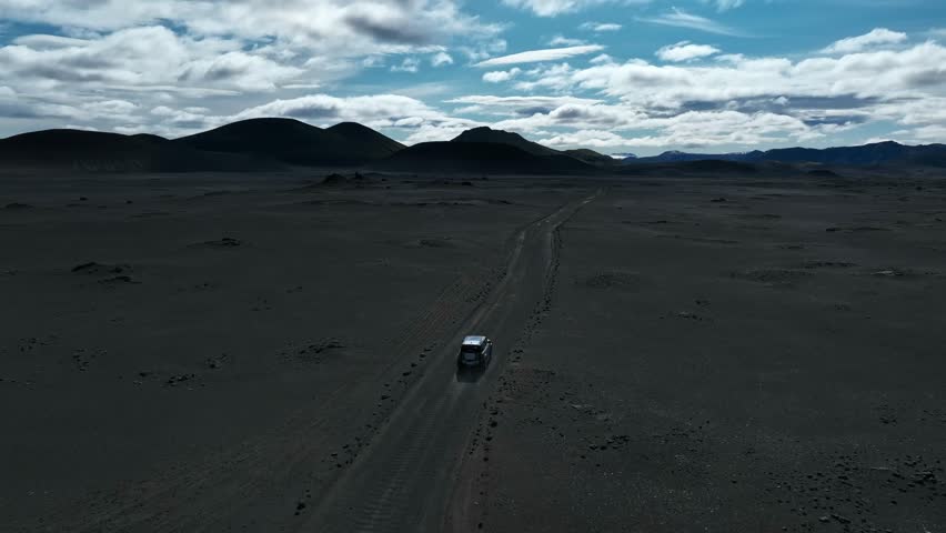 A lone 4x4 drives across Iceland’s black volcanic desert under bright clouds, crossing the remote Fjallabak wilderness with mountains on the horizon.