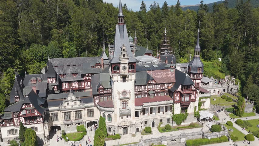 Drone orbiting around Peles Castle, showing its roofs, towers, and forested surroundings.