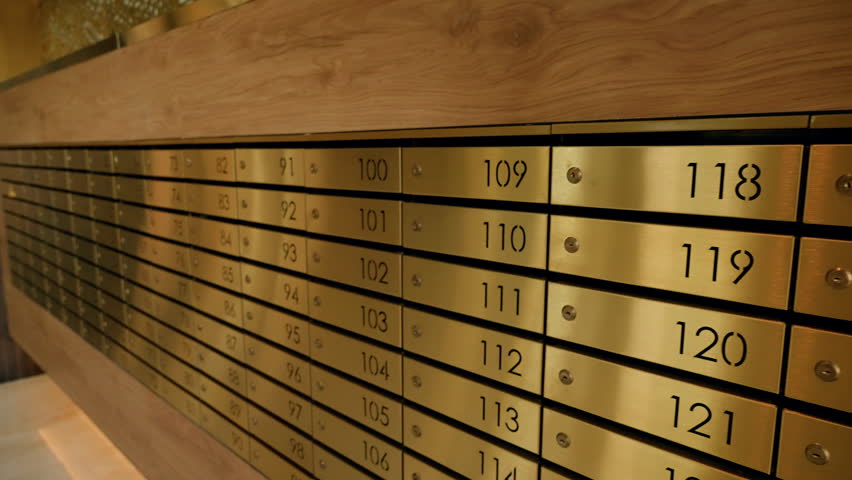 Warm-lit row of brass numbered mailboxes built into a wooden wall in a residential lobby, with horizontal compartments and sequential numbering for postal delivery and storage