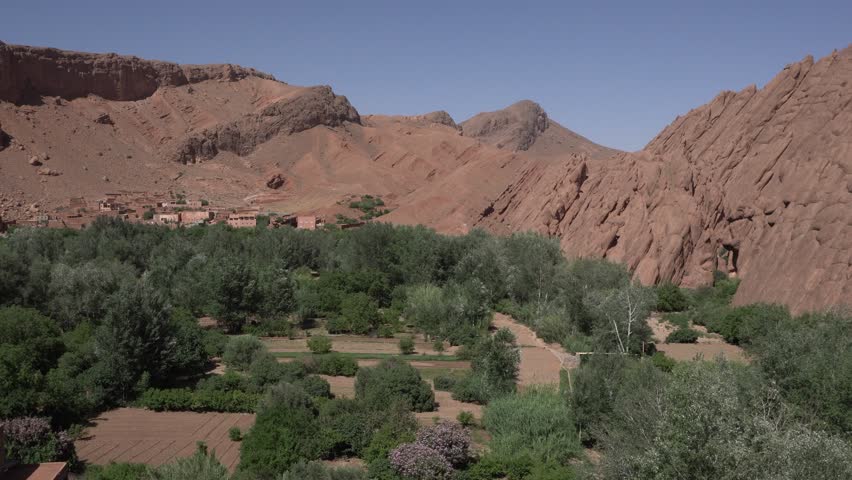 Several desert mountains under a magnificent blue sky. In the valley, you can see cultivated fields with abundant vegetation and some structures.