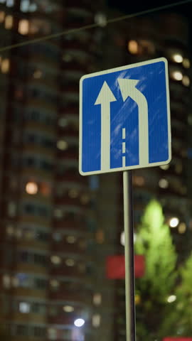 Nighttime urban scene with a blue traffic sign showing a straight-ahead lane and a right lane merging left, set against high-rise apartments and soft city lights.