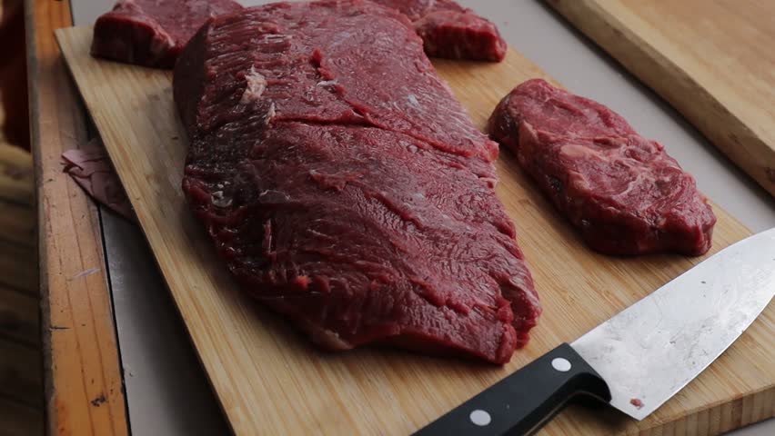 Close-up of gloved hands slicing a piece of raw steak with a sharp knife on a wooden cutting board, prepared for cooking or portioning.