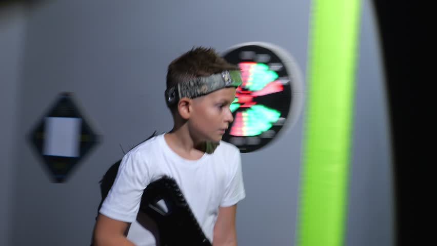 A cheerful teenage boy with a laser pistol is having fun in a dark laser tag arena, he runs around the arena and aims at the enemy. A colorful laser tag arena with neon stripes on the walls.