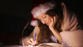 At night, cheerful woman wearing santa hat is lying in bed, smiling as she writes warm christmas letter to santa claus or cherished loved one, illuminated by the soft glow of a small lamp - Powered by Shutterstock - Get 15% off with code: PIKWIZARD15