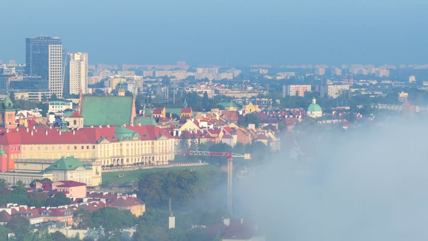 Aerial view of Warsaw’s Royal Castle and Old Town rooftops emerging above dense morning fog drifting over Vistula River, with modern city skyline in distance. Wide shot.