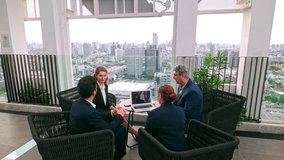 Two business professionals shake hands outdoors on a rooftop, while colleagues sit nearby with a laptop. The group engages in a collaborative meeting with a city skyline in the background. 4k video - Powered by Shutterstock - Get 15% off with code: PIKWIZARD15
