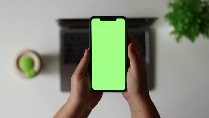 Woman sit use digital tech cell phone, green screen mock up. Hand scroll iphone web site, swipe like photos. Girl hold mobile closeup. Chroma key page. Cozy office background. Online app copy space.
