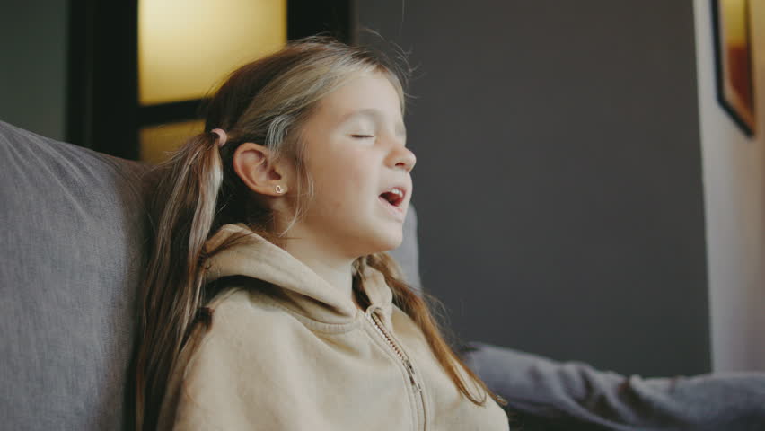 A sad girl sits on a couch in a softly lit room, her emotions visible as she expresses herself through her facial reaction and body language.