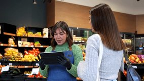 Friendly female grocery store clerk wearing an apron and gloves using a digital tablet to assist a woman with her shopping, providing excellent customer service in the produce section - Powered by Shutterstock - Get 15% off with code: PIKWIZARD15