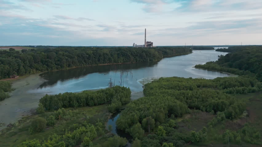 Aerial of lake inlet bordered by forested slopes with calm water reflecting muted sky, processing industrial plant on horizon