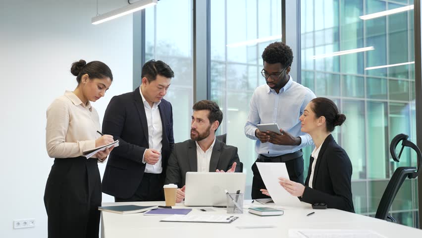 Male boss with group of diverse employees discussing work issues using laptop at meeting in office. Manager explains details of project development to subordinates, gives tasks, engaging in teamwork