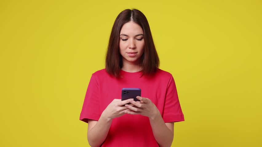 Happy excited joyful young woman wear pink t-shirt casual clothes hold use pointing finger on mobile cell phone just found out great big win news isolated on plain yellow background. Lifestyle concept