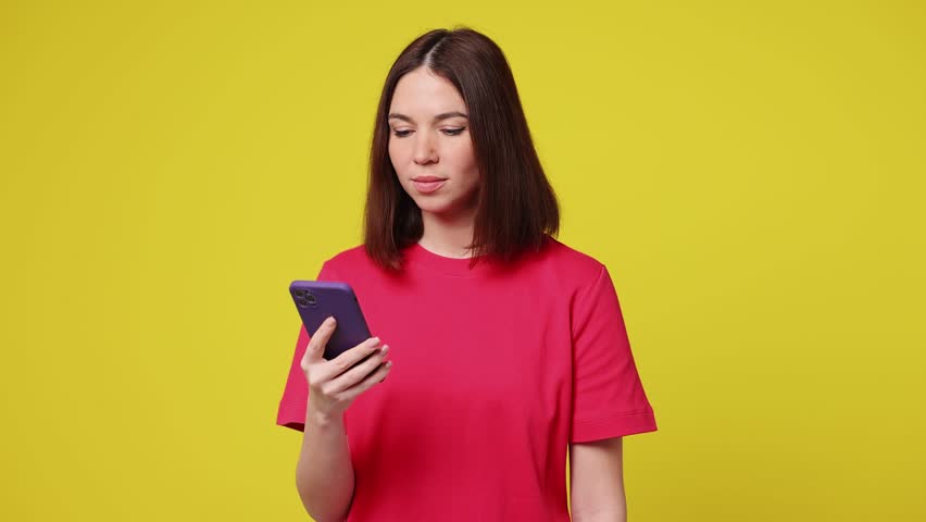 Excited happy joyful young woman she wear pink t-shirt casual clothes use mobile cell phone hold win fan of cash money in dollar banknotes isolated on plain yellow color background. Lifestyle concept