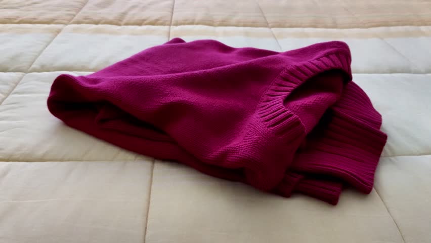 Ribbed magenta sweater draped softly across a pale grid-stitch quilt, highlighting tactile fabric contrast and cozy layered textures in a calm, minimal home setting