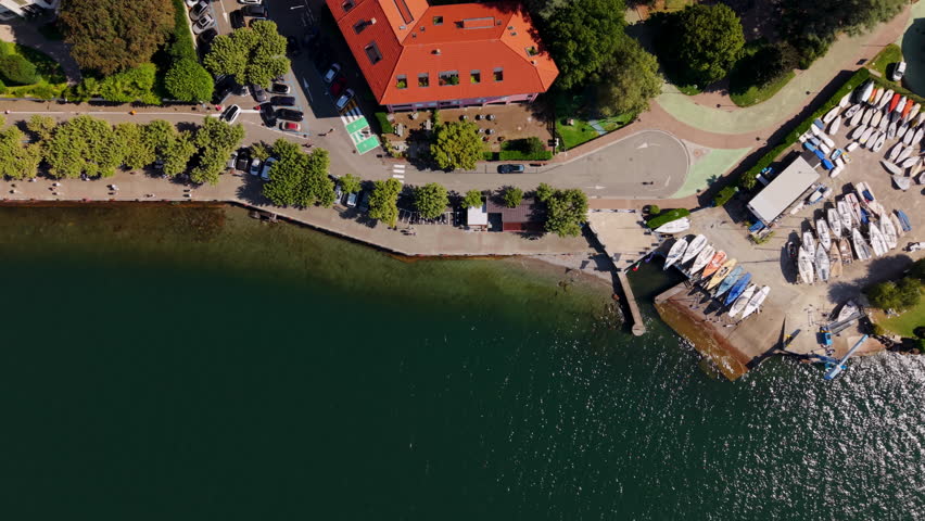Top-down drone panning over a lakeside road with trees, houses, parked cars, and the waterfront below.