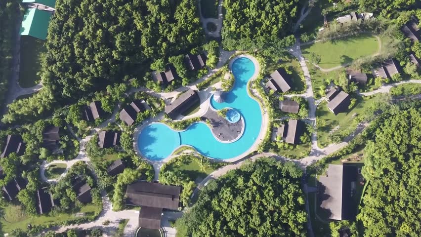 Aerial drone shot showcasing a lush tropical resort with winding paths, dark-roofed bungalows, and a vibrant blue, multi-level swimming pool.