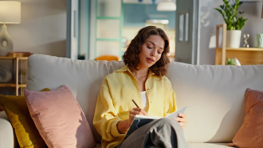 Inspired girl writing in copybook sitting comfortable home couch closeup. Smiling curly woman thinking creative ideas making notes in notebook. Relaxed brunette keeping diary in living room apartment.