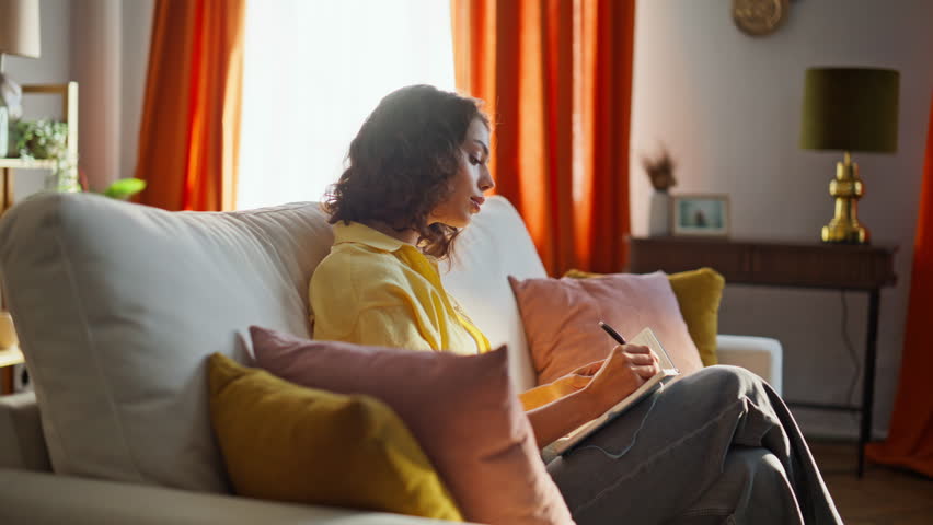 Home lady writing planner relaxing at cozy apartment couch. Smiling beautiful brunette thinking creative ideas making notes in notebook. Relaxed woman writer holding pen creating novel in living room.