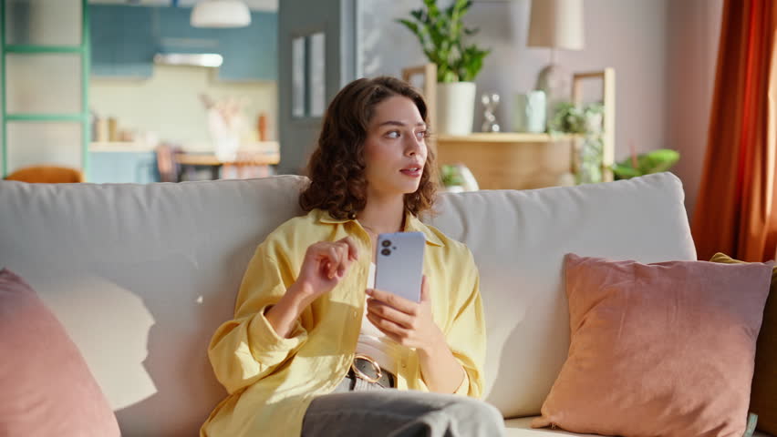 Relaxed girl scrolling cellphone at comfortable couch. Smiling curly woman watching news feed on smartphone sitting cozy home sofa alone. Calm brunette student watching social media content on phone.