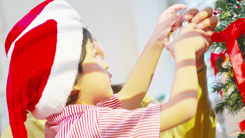 Asian Festive child wearing santa hat decorates christmas tree with parent helping gentle joyful scene child wearing red santa hat hangs ornament christmas tree joyful holiday family moment child
