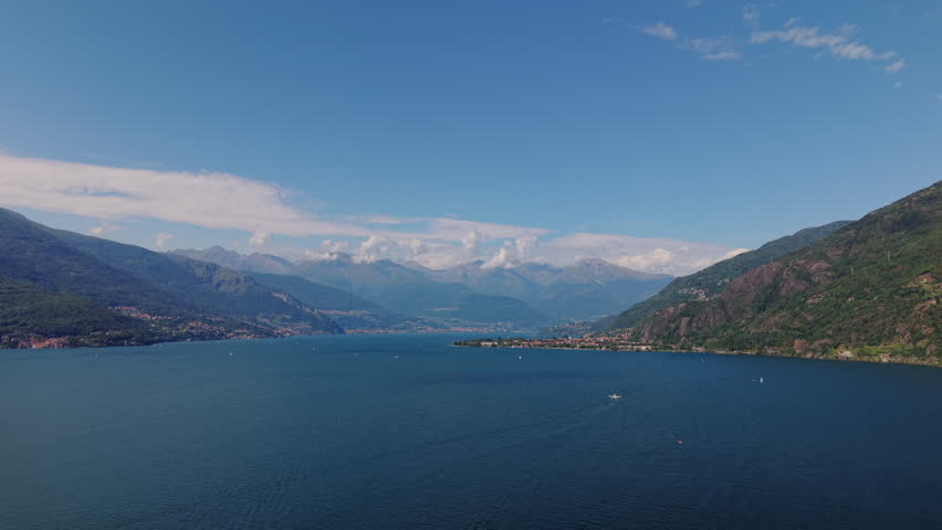 Drone advances over Lake Como, revealing the wide water surface, small distant boats, layered mountains and soft clouds under clear summer light.