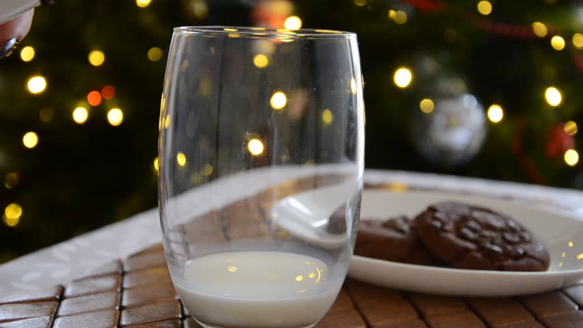 Milk being poured into a clear glass in front of chocolate cookies and a glowing Christmas tree, creating a cozy festive atmosphere perfect for holiday food scenes, seasonal storytelling, and warm win