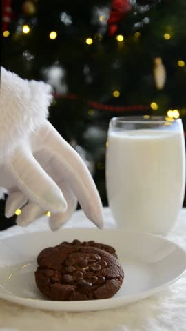Santa’s gloved hand taking a bitten chocolate cookie and lifting a glass of milk in front of a glowing Christmas tree, capturing warm festive traditions and cozy holiday atmosphere