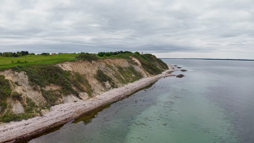 Drone aerial footage capturing rocky coastal cliffs with green vegetation along the shoreline and calm sea waters in Denmark.